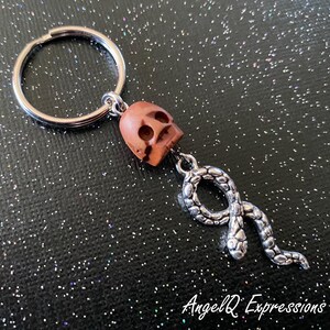 May include: A silver key chain with a wooden skull charm and a silver snake charm. The skull is brown and has a detailed design. The snake is curled up and has a textured surface.