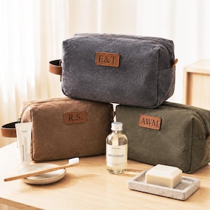 Puede incluir: Tres neceseres rectangulares de lona en gris carbón, marrón y verde oliva, cada uno con un parche de cuero marrón con las iniciales E&T, R.S. y AWM grabadas. Los bolsos tienen asas laterales y cremalleras, acompañados de artículos de aseo.