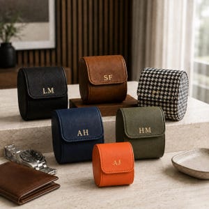 May include: A collection of six cylindrical watch cases in various colours including black, brown, navy blue, olive green, orange, and a black and white houndstooth pattern. Each case features a flap closure with gold embossed initials.