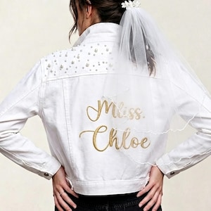 May include: White denim jacket with pearl embellishments on the shoulders and gold embroidered text that reads "Miss Chloe". A white veil is attached to the jacket. Bridal wear.
