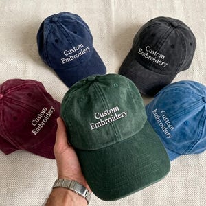 Può includere: Cinque cappellini da baseball in vari colori: blu navy, nero, bordeaux, verde e blu. Ogni cappellino presenta il ricamo bianco "Custom Embroidery". I cappellini sono disposti su una superficie chiara e strutturata.