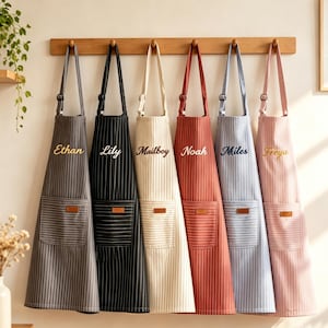 May include: Six striped aprons hanging on a wooden rack, each featuring a front pocket with a leather-like patch and a personalized name embroidered in cursive script: Ethan, Lily, Mailboy, Noah, Miles, and Freya, in gray, black, cream, red, blue, and pink.