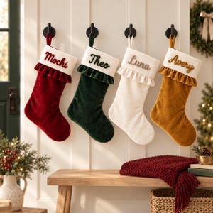May include: Four velvet Christmas stockings hanging on black wall hooks. Each stocking features a white cuff with a ruffled edge and embroidered names: Mochi in red, Theo in dark green, Luna in tan, and Avery in brown. The stockings are red, green, cream, and mustard yellow.