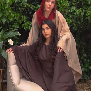 May include: Two women wearing long robes, one in brown and one in beige with a red headscarf. The woman in brown is sitting on a light beige armchair.