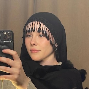 Può includere: Un hijab nero con una frangia di perline e un bordo in pelliccia nera. L'hijab è indossato da una persona con la pelle chiara e i capelli castano chiaro.