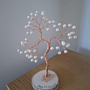 May include: A decorative wire tree sculpture crafted from copper-coloured wire with white pearl-like beads at the ends of the branches. The tree is mounted on a circular wooden slice base with the text Happy 4th Anniversary written in black ink.