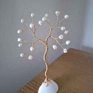 May include: A decorative tree sculpture crafted from twisted gold-coloured wire, featuring small white pearls attached to the ends of the branches. The base of the tree is formed by a single white seashell, providing a stable foundation for the structure.