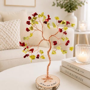 May include: A decorative wire tree sculpture featuring twisted copper-colored wire branches adorned with small, polished red and light green gemstone chips. The tree is mounted on a circular wooden base with the text 35 Years and 35 More!.