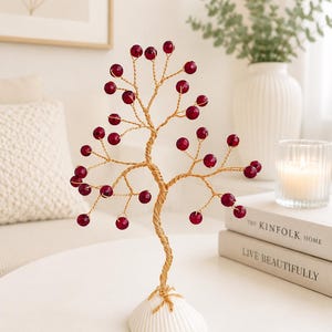 May include: A decorative wire tree sculpture featuring a twisted gold-tone metal trunk and branches adorned with small, round, deep red beads. The tree is mounted on a white, ribbed, semi-circular seashell base, creating a balanced, upright structure.