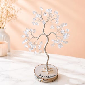 May include: A decorative wire tree sculpture featuring clear crystal-like stones on silver-toned branches. The tree is mounted on a circular wooden base with the text "Happy 15th Anniversary" written in black ink on the front edge of the wood.