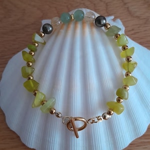 May include: A beaded bracelet featuring light green and gold-colored stones. The bracelet has a gold-colored clasp and is displayed on a white seashell. The stones are irregular in shape and are interspersed with small gold beads.