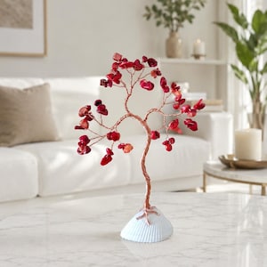 May include: A decorative tree sculpture featuring a twisted copper wire trunk and branches adorned with numerous small, polished red gemstone chips. The tree is mounted on a white, ribbed seashell base that serves as a stable foundation.