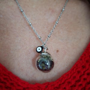 Puede incluir: Un collar plateado con un pequeño colgante de orbe de vidrio transparente lleno de flores y vegetación secas. Un pequeño amuleto blanco y negro cuelga sobre el orbe. El collar se muestra sobre un suéter rojo tejido.