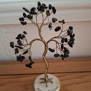 May include: A handcrafted wire tree sculpture with black gemstone leaves. The tree trunk and branches are made of twisted gold-colored wire. The tree is mounted on a small, round wooden base with the words "Happy 8th Anniversary" written on it.