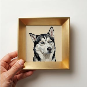 Könnte beinhalten: Ein quadratisches gerahmtes Kunstwerk mit einer stilisierten Illustration eines Siberian Husky mit schwarzem, weißem und grauem Fell. Der Hund hat hellblaue Augen und blickt nach rechts. Der Rahmen besteht aus gebürstetem goldfarbenem Metall.