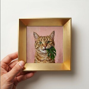 Könnte beinhalten: Ein quadratisches gerahmtes Gemälde, das eine braune getigerte Katze mit grünen Augen zeigt, die ein grünes Monstera-Blatt im Maul hält. Die Katze ist vor einem einfarbigen, altrosa Hintergrund dargestellt. Das Bild steckt in einem Goldrahmen.