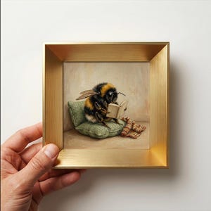 Könnte beinhalten: Ein quadratisch gerahmter Kunstdruck mit einem Gemälde einer flauschigen Hummel, die auf einem kleinen grünen Strickkissen sitzt und ein offenes Buch liest. Neben der Hummel liegt eine gefaltete, braun-weiß gemusterte Strickdecke. Der Rahmen ist gold.