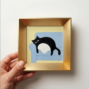 Könnte beinhalten: Ein quadratisches Gemälde in einem goldfarbenen Metallrahmen zeigt eine schwarze Katze mit weißem Bauch, die auf einem hellblauen Sofa vor einer blassgelben Wand liegt. Die Katze hat große Augen und liegt horizontal auf dem blauen Möbelstück.