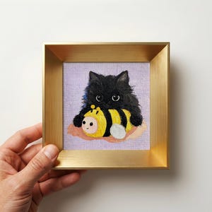 Könnte beinhalten: Ein kleines quadratisches Gemälde in einem metallisch goldenen Rahmen zeigt eine flauschige schwarze Katze, die ein gelb-schwarz gestreiftes Bienenspielzeug umarmt. Die Katze hat große dunkle Augen vor einem hellvioletten Hintergrund mit einem rosa Fleck.