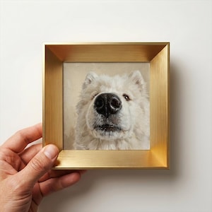 Könnte beinhalten: Ein quadratisch gerahmtes Gemälde mit einem Nahporträt eines weißen Samojeden-Hundes mit einer markanten schwarzen Nase. Das Kunstwerk ist in einem gebürsteten Goldmetallrahmen mit tiefem, abgeschrägtem Profil gefasst, das das weiße Fell betont.