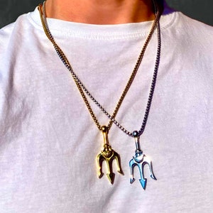 Puede incluir: Dos collares de oro y plata con colgantes de tridente. Los collares están superpuestos en el cuello de una persona.