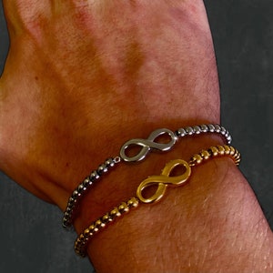 Op de afbeelding: Twee armbanden met kralen en oneindigheidssymbolen. Eén armband is zilverkleurig, de andere goudkleurig. De armbanden zijn gemaakt van kleine, ronde kralen. De oneindigheidssymbolen zijn het centrale ontwerpelement.