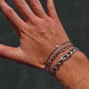 Könnte beinhalten: Drei silberne Kettenarmbänder, die am Handgelenk einer Person übereinander liegen. Die Armbänder haben unterschiedliche Breiten und Stile.