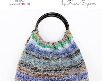Puede incluir: Una bolsa de punto de rayas de colores con un asa de madera negra. La bolsa está hecha con un hilo grueso y tiene una forma relajada y holgada. El texto "CLASSIC CHIC KNIT BAG by Kari Capone" está impreso encima de la bolsa.