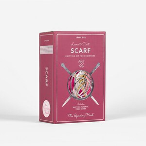 Op de afbeelding: Een bordeauxrode doos om te leren een sjaal te breien. De doos bevat de tekst "Learn to Knit SCARF Knitting Kit for Beginners" en bevat een video-tutorialserie over breien. Het logo The Spinning Hand is zichtbaar.
