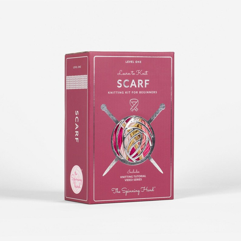 Op de afbeelding: Een bordeauxrode doos om te leren een sjaal te breien. De doos bevat de tekst "Learn to Knit SCARF Knitting Kit for Beginners" en bevat een video-tutorialserie over breien. Het logo The Spinning Hand is zichtbaar.