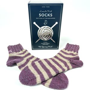 May include: Hand-knitted socks in purple and cream stripes, with a knitting kit box. The box reads "Learn to Knit SOCKS" and includes a knitting tutorial video series. The socks are a beginner's knitting project.