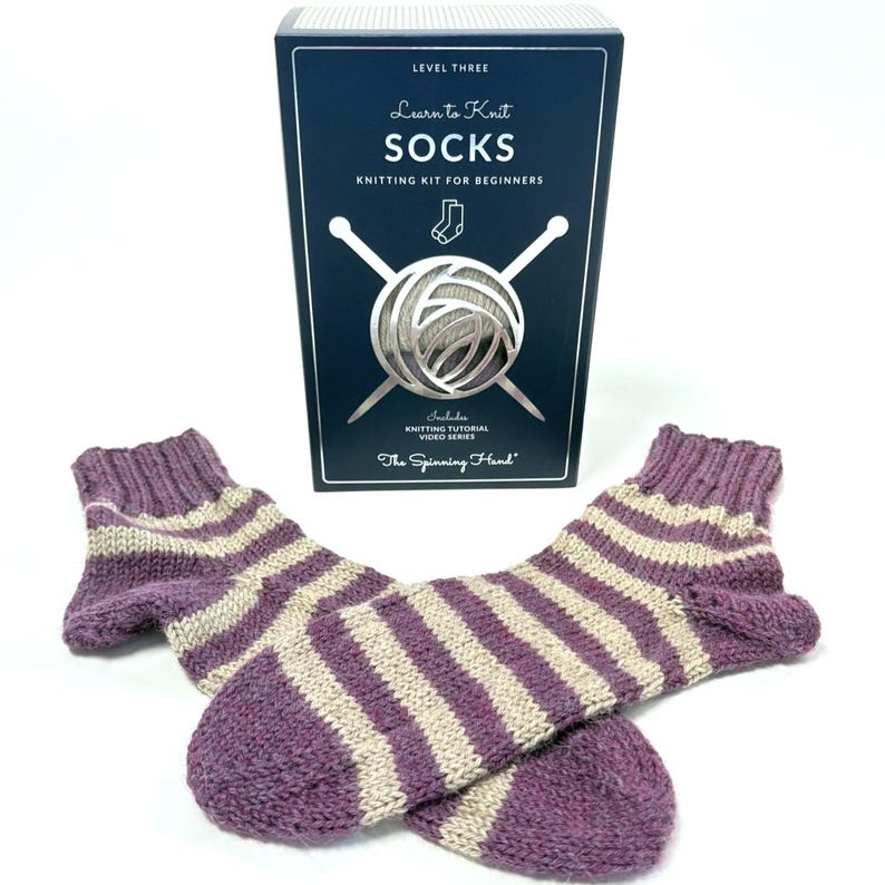 May include: Hand-knitted socks in purple and cream stripes, with a knitting kit box. The box reads "Learn to Knit SOCKS" and includes a knitting tutorial video series. The socks are a beginner's knitting project.