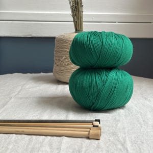 Peut inclure: Deux pelotes de laine vert vif empilées. Derrière, une grosse bobine de fil beige et un bouquet d'herbes séchées. Des aiguilles à tricoter sont au premier plan. Fournitures de tricot.