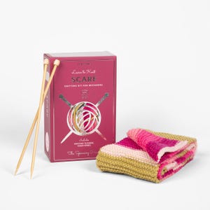 Op de afbeelding: Een breipakket voor beginners met een sjaal, breinaalden en garen. De magenta doos draagt de tekst "Learn to Knit SCARF" en bevat een video-tutorialserie. De gevouwen sjaal toont roze, witte en gele strepen. De breinaalden zijn van hout.