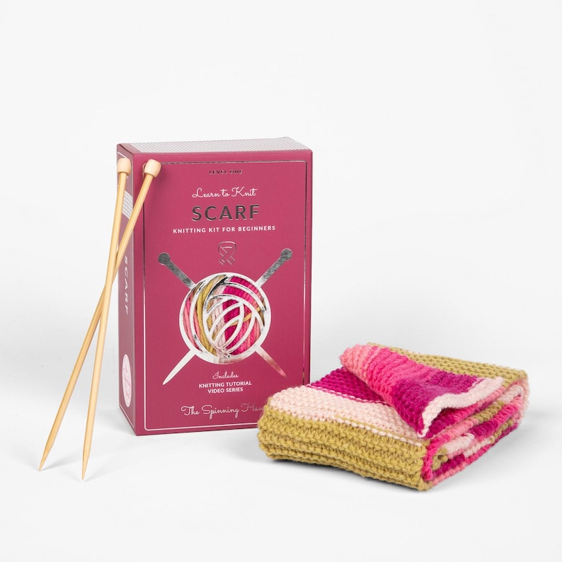 Op de afbeelding: Een breipakket voor beginners met een sjaal, breinaalden en garen. De magenta doos draagt de tekst "Learn to Knit SCARF" en bevat een video-tutorialserie. De gevouwen sjaal toont roze, witte en gele strepen. De breinaalden zijn van hout.