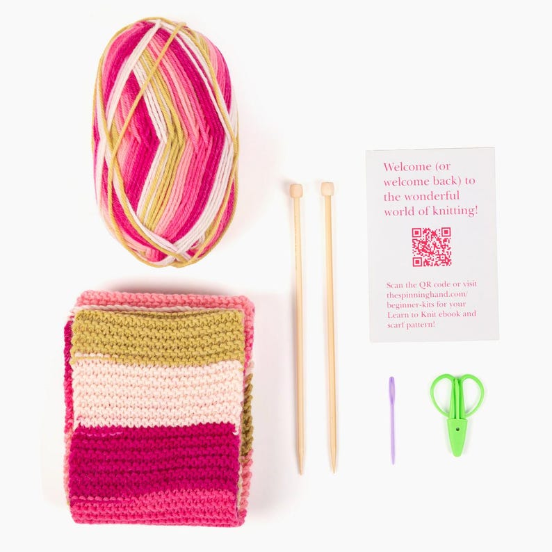 Op de afbeelding: Een breipakket met een bol roze, witte en gele wol, een gebreide sjaal, twee houten breinaalden, een groene schaar, een paarse naald en een kaart met tekst: "Welcome to the wonderful world of knitting!"