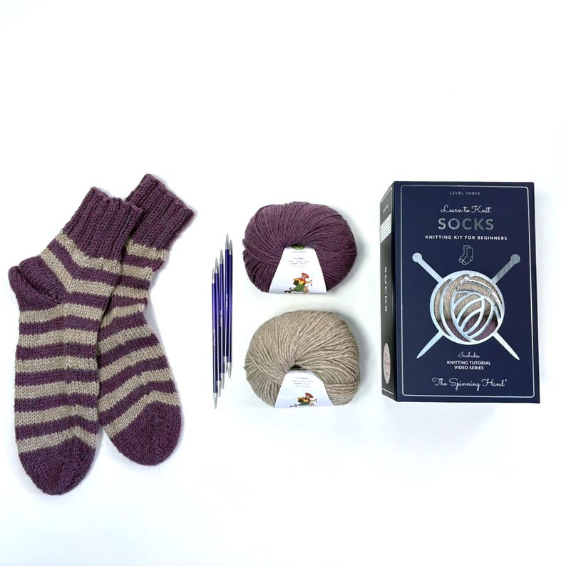 May include: A knitting kit for beginners, including a pair of purple and beige striped socks, two balls of yarn, knitting needles, and a box set. The box reads "Learn to Knit SOCKS Knitting Kit for Beginners" and "The Spinning Hand".