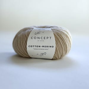 May include: A ball of beige yarn with a white label wrapped around it. The label reads "CONCEPT BY KATIA COTTON-MERINO Merino Extrafine Wool". The yarn is a light beige color and appears soft and textured, suitable for knitting or crochet projects.