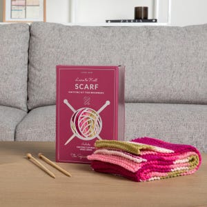 Op de afbeelding: Een breipakket voor beginners om een sjaal te leren breien. De set bevat een doos met de tekst "Learn to Knit SCARF" en een afbeelding van wol en breinaalden. Een gevouwen sjaal in roze, rode en groene tinten is ook zichtbaar.