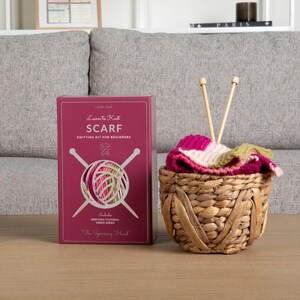 Op de afbeelding: Een breipakket voor beginners met een bordeauxrode doos met de tekst "Learn to Knit SCARF". De set bevat een mand met een gedeeltelijk gebreide sjaal in roze, wit en groen, samen met twee houten breinaalden. De tekst "The Spinning Hand" is ook zichtbaar.