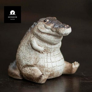 May include: A white ceramic figurine of a crocodile with a wide, toothy grin. The crocodile is sitting with its legs crossed and has a rounded, plump body.