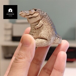 May include: A small, hand-painted ceramic alligator figurine in a light brown and dark brown colour scheme. The alligator is sitting with its legs tucked under its body and has a friendly expression.