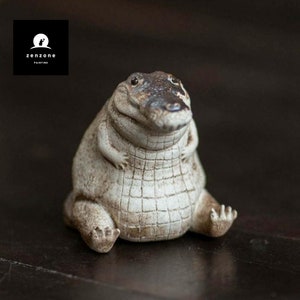 May include: A ceramic figurine of a crocodile with a white and brown colour scheme. The crocodile is sitting with its legs crossed and has a wide, toothy grin.