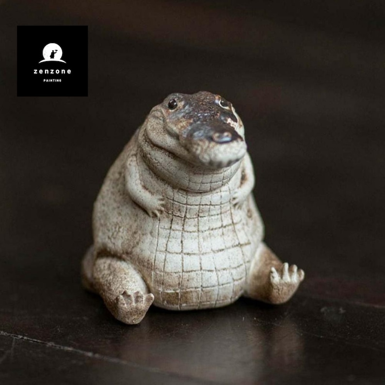 May include: A ceramic figurine of a crocodile with a white and brown colour scheme. The crocodile is sitting with its legs crossed and has a wide, toothy grin.