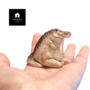 May include: A small, brown and tan alligator figurine with a wide, toothy grin. The alligator is sitting on an open hand.