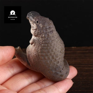 May include: A small, brown ceramic sculpture of a crocodile sitting on its hind legs with its head tilted up. The sculpture has a textured surface that resembles crocodile scales.
