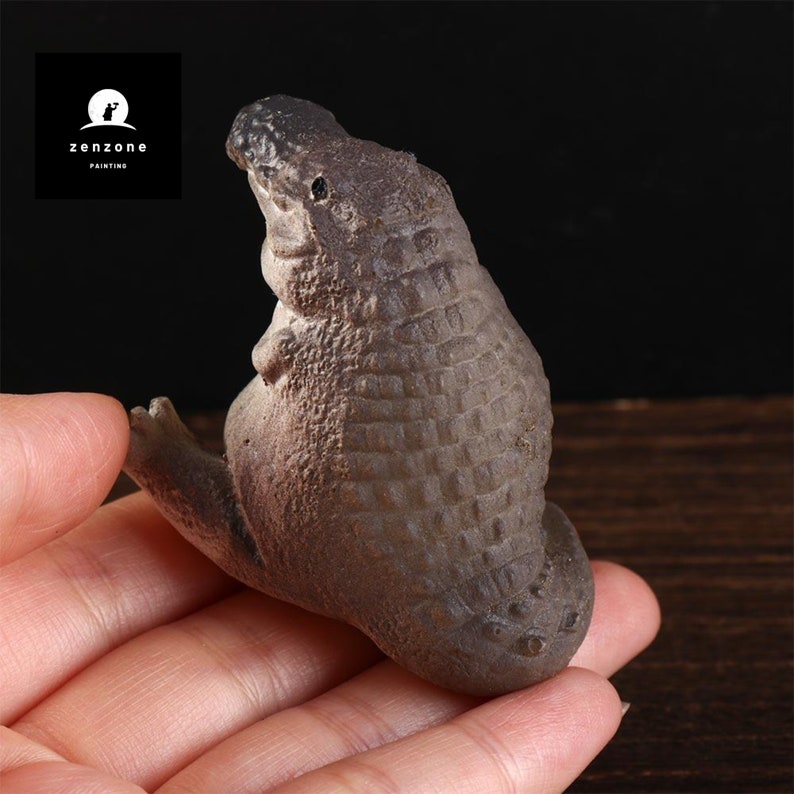 May include: A small, brown ceramic sculpture of a crocodile sitting on its hind legs with its head tilted up. The sculpture has a textured surface that resembles crocodile scales.