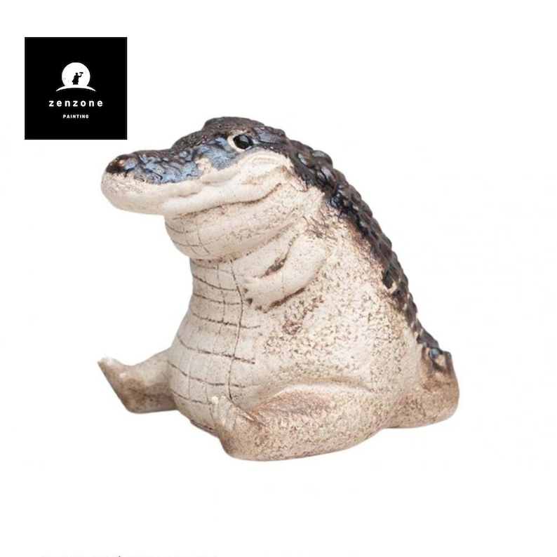 May include: A ceramic figurine of a smiling alligator sitting with its legs crossed. The alligator is painted in shades of brown and white.