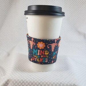 Peut inclure: Un gobelet à café en papier blanc avec un couvercle noir, entouré d'une manchette en tissu bleu marine. La manchette présente un motif coloré avec le texte "MIND YOUR OWN UTERUS" et des illustrations de fleurs et d'organes reproducteurs.