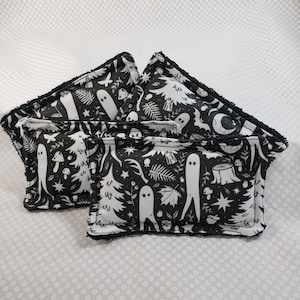 May include: Four black and white decorative pillows. The pillows feature a repeating pattern of white ghosts, trees, mushrooms, and other forest elements on a black background. The edges of the pillows are trimmed with black fringe.